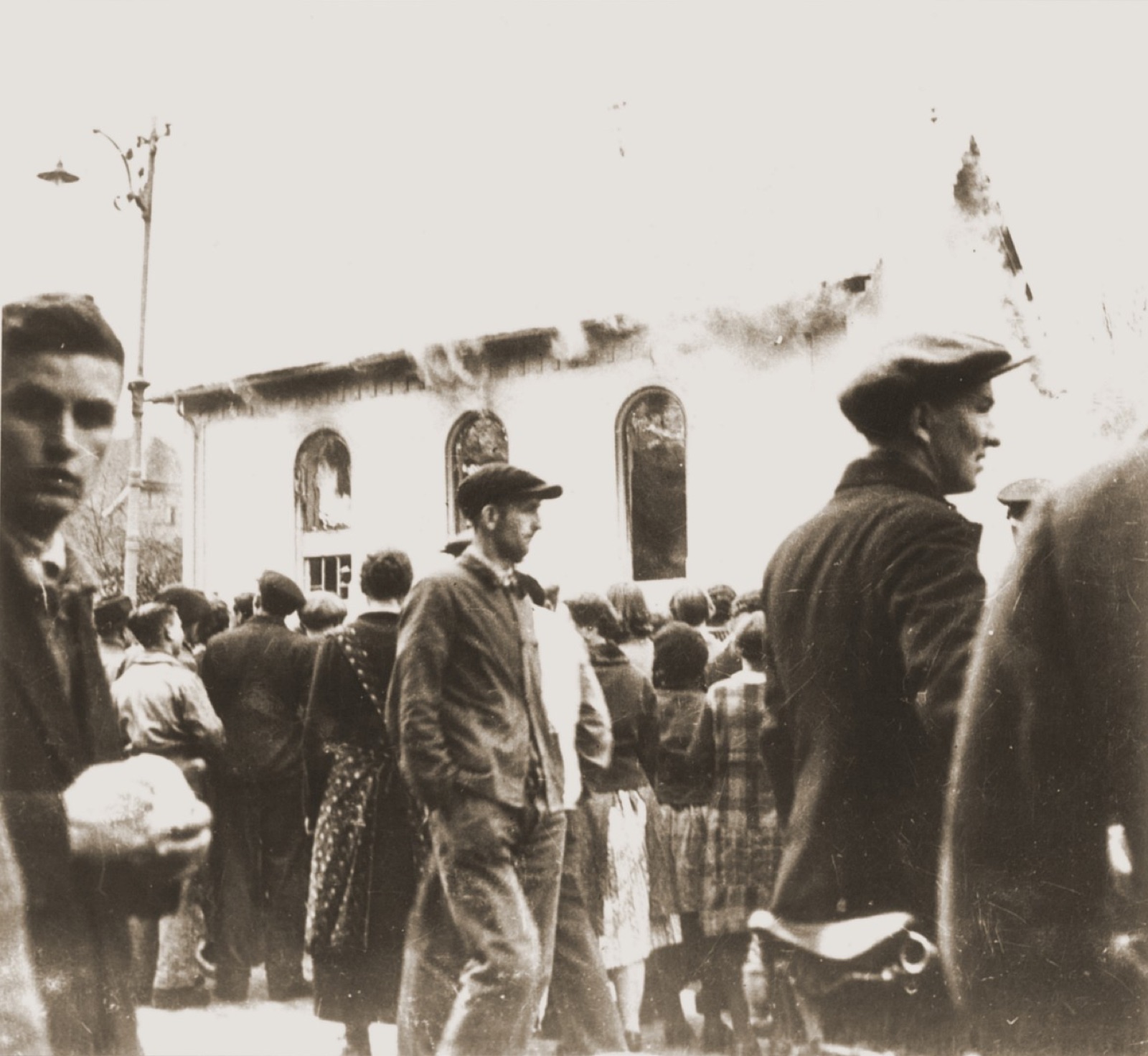 A line of townspeople in coats and hats standing in a village street, turned toward a building that is burning; smoke rises above the roofline and a fire crew works off to one side.