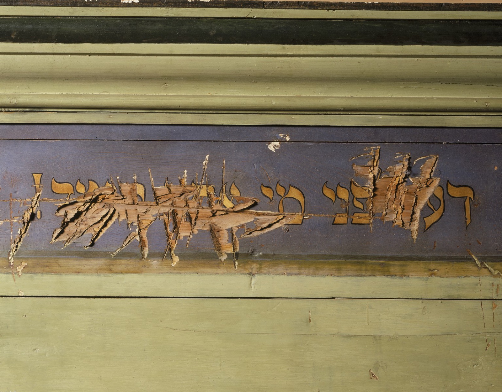 A painted lintel photographed on its own, its gilded Hebrew inscription hacked away across much of its length.