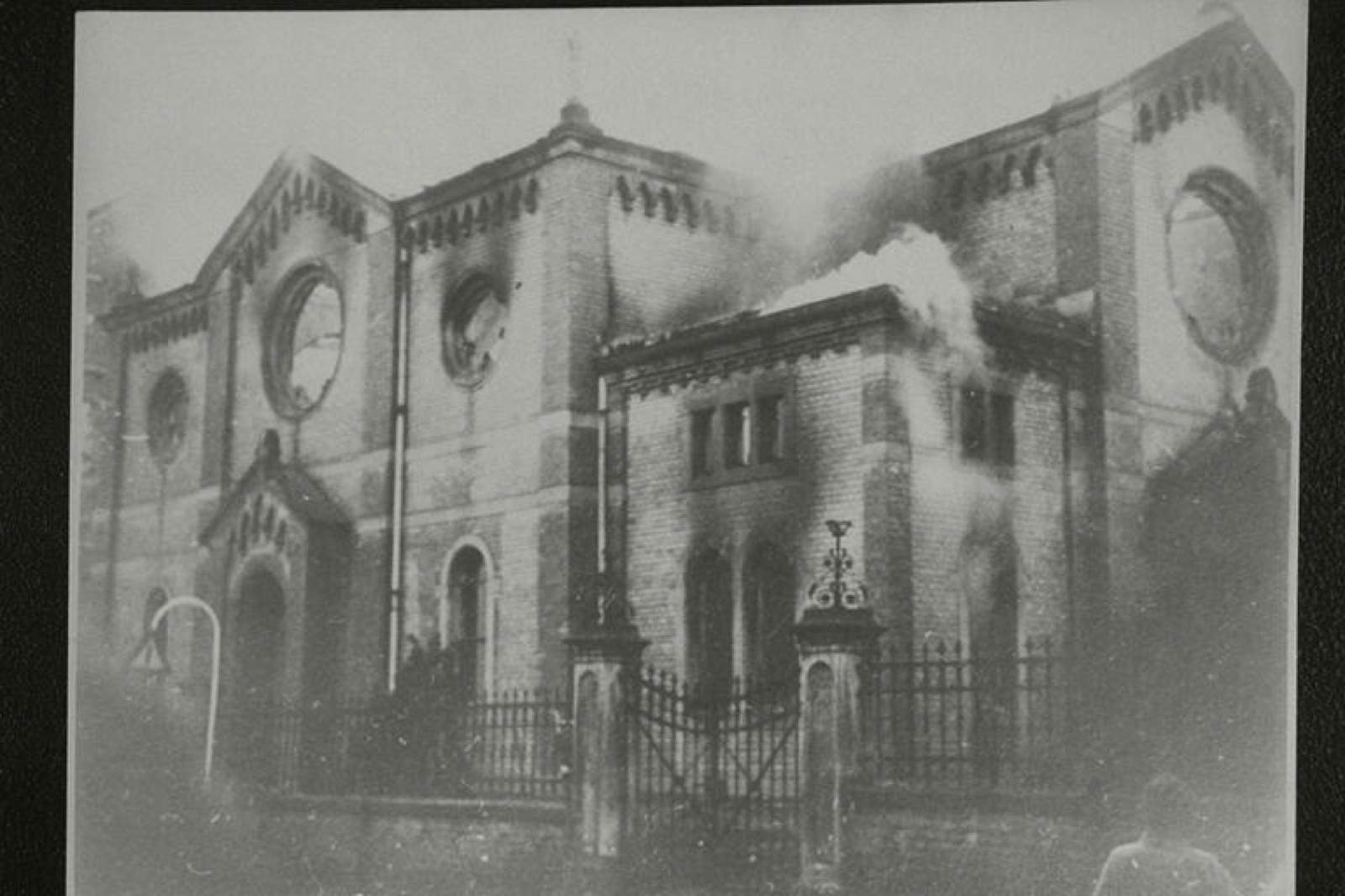 A synagogue on fire at night, flames breaking through the roof and smoke rising above the surrounding rooftops.