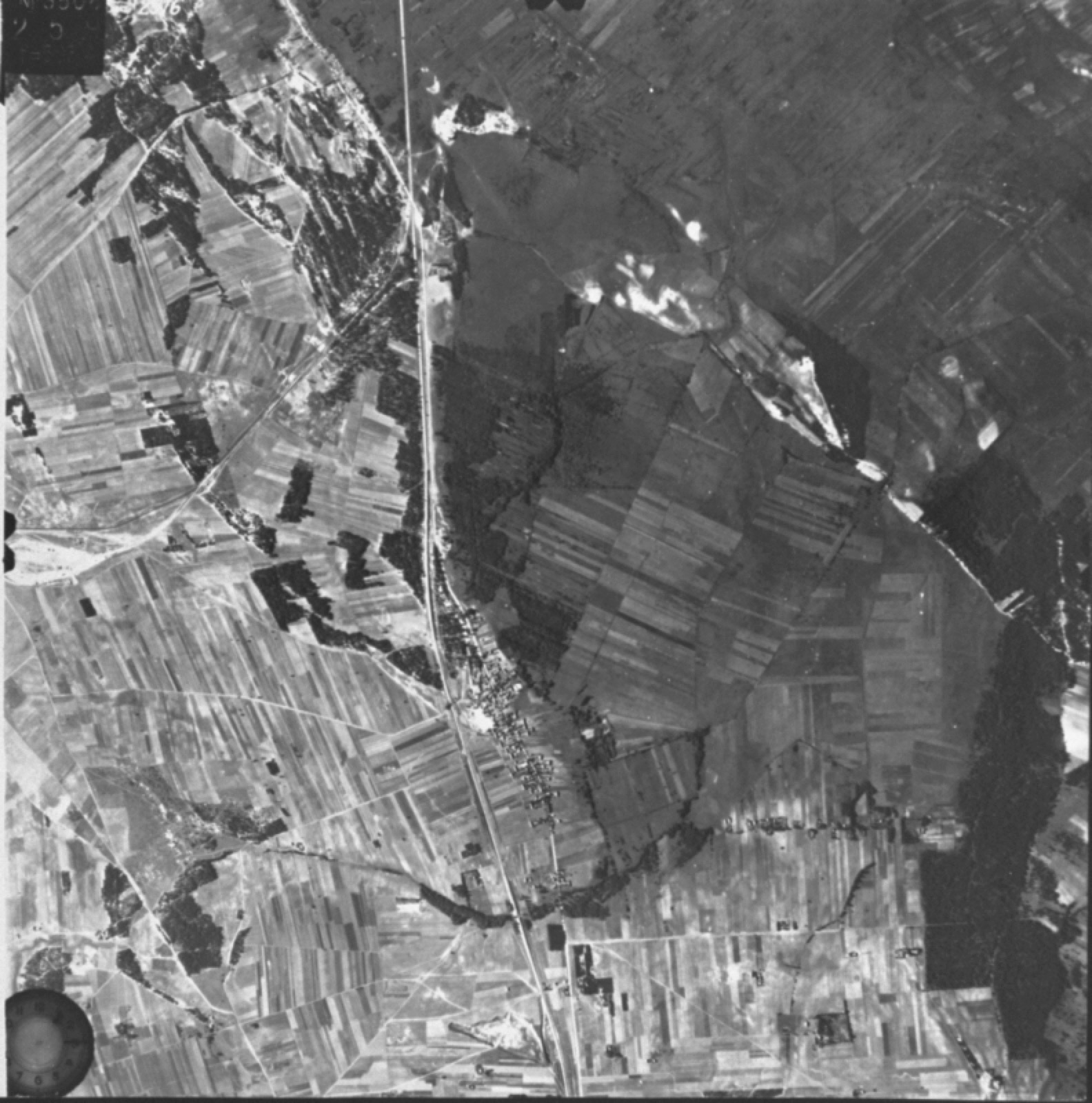 Low-contrast aerial photograph of open ground and tree lines, with the faint outline of a cleared rectangular area and a rail spur.