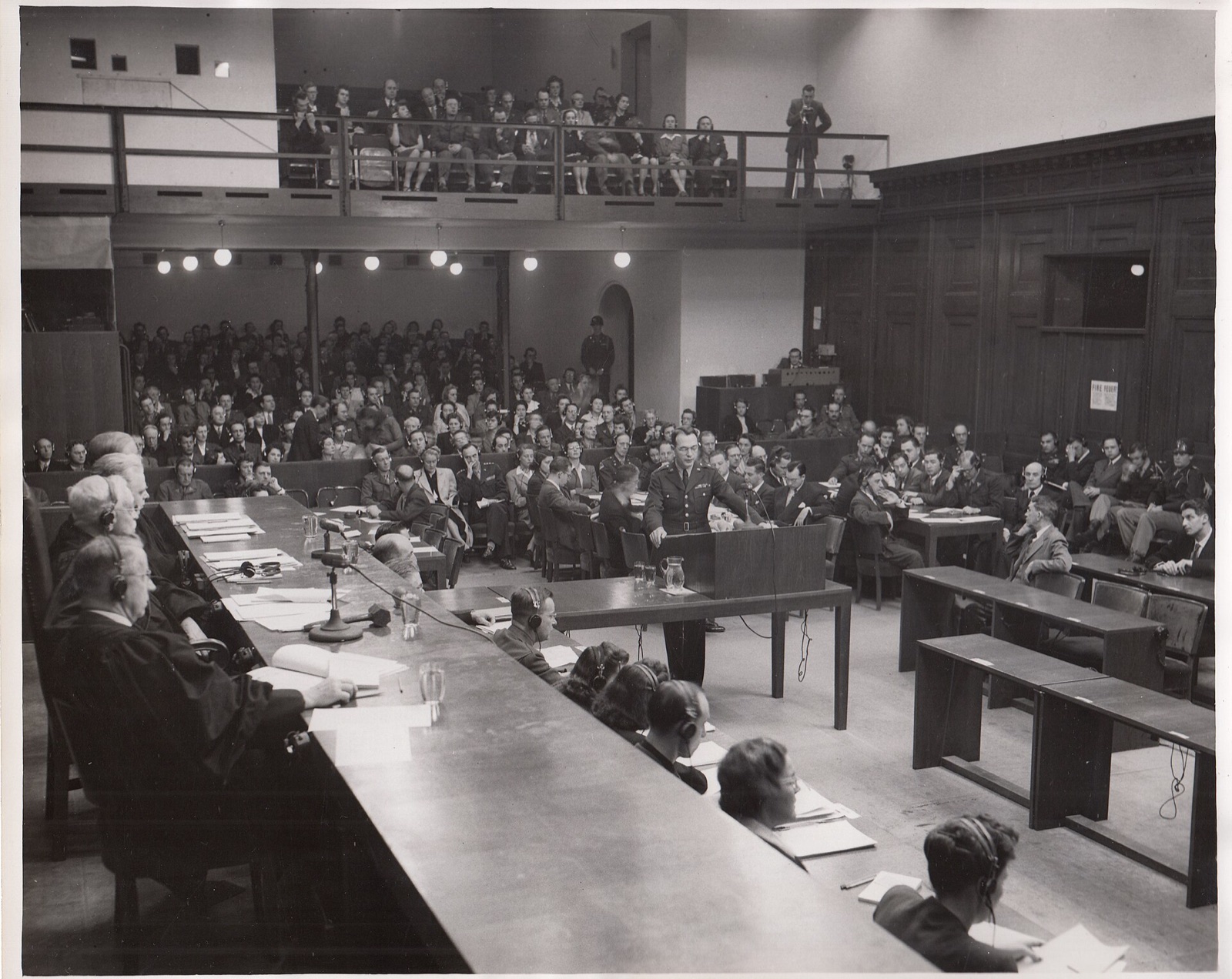 A uniformed officer stands at a lectern in a crowded courtroom, papers in one hand, speaking toward the bench.