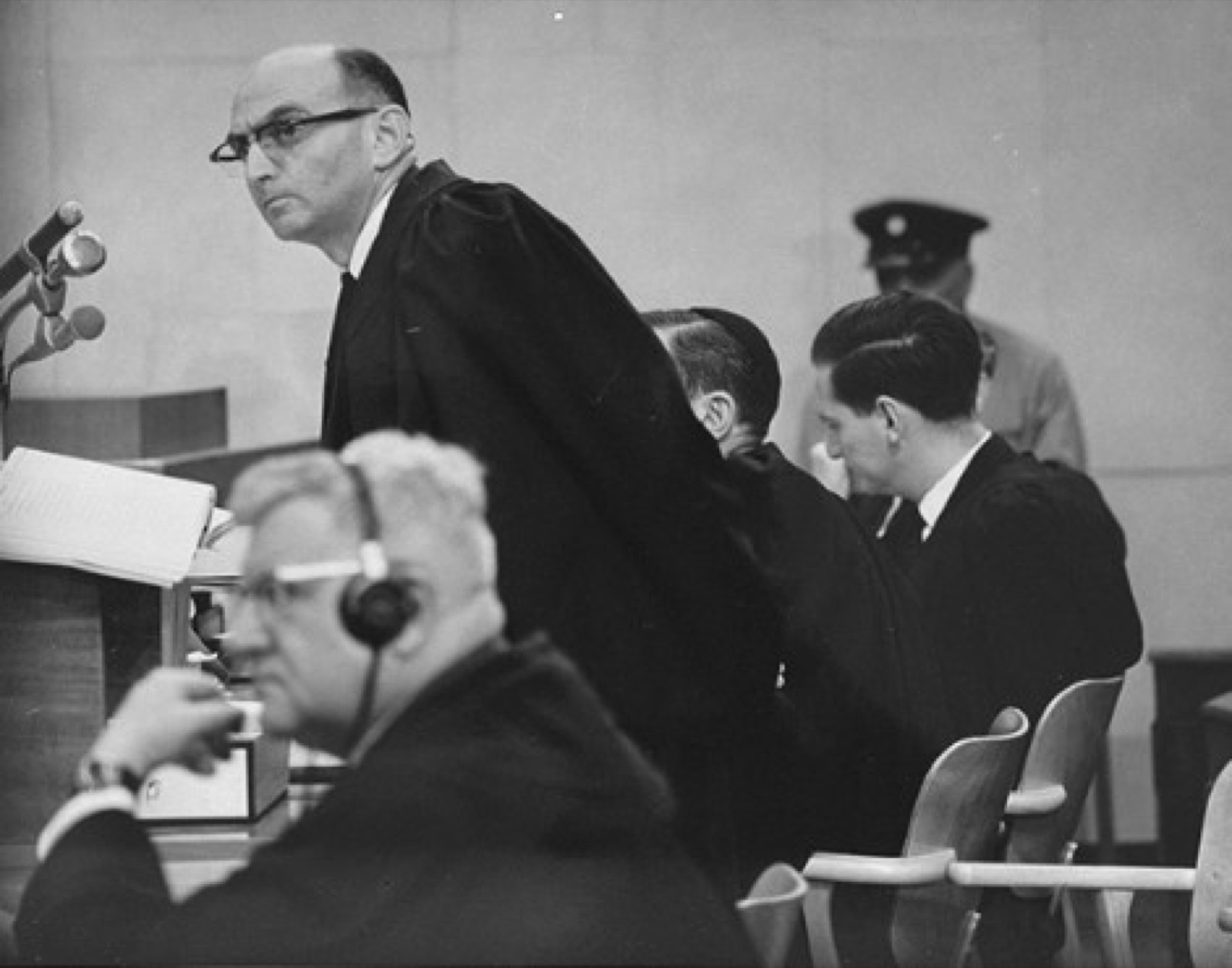 Two men in a courtroom, one seated at a table wearing headphones and the other standing beside him in a dark suit, files and microphones on the table between them.