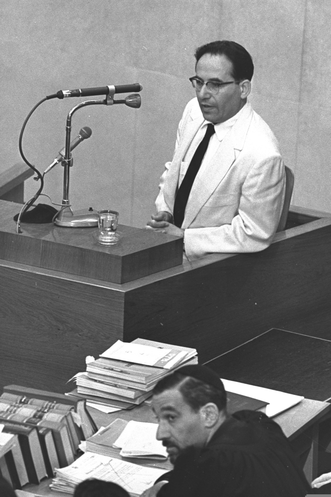 A man in a light suit stands at a witness stand with a microphone in front of him, one hand raised slightly, his face drawn.