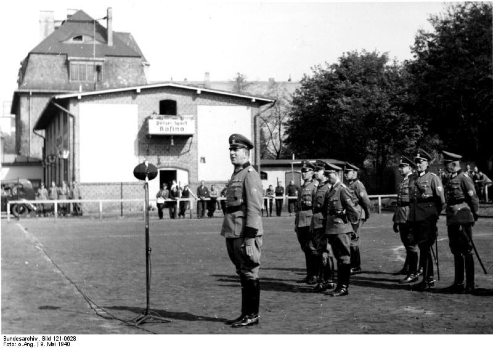 Ranks of German policemen in steel helmets marching across a parade ground in daylight, rifles slung, several of them carrying suitcases.
